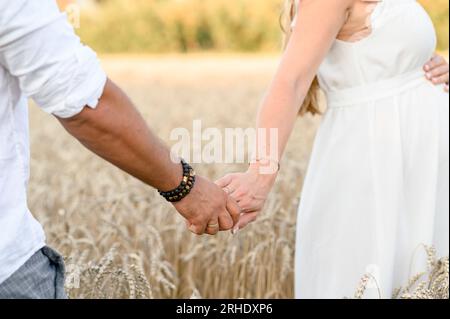 Seitenansicht einer anonymen schwangeren Frau in weißem Kleid, die den Bauch berührt und steht, während sie die Hand eines nicht erkennbaren Mannes im Weizenfeld in s hält Stockfoto