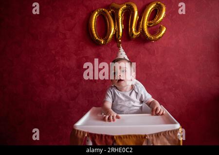 Portrait eines einjährigen Jungen mit festlichem Hut sitzt auf einem Kinderstuhl mit Stofffahnen. Kind auf isoliertem roten Hintergrund mit goldenen Luftballons aus Folie Stockfoto