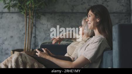 Seitenansicht einer glücklichen erwachsenen Frau mit ihrer Seniormutter, die zu Hause Spaß beim Spielen von Konsolenvideospielen hat. Zwei Generationen haben Spaß Stockfoto