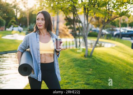 Selbstbewusste junge Frau, die morgens Kaffee im Park mit Yoga-Matte genießt. Halten der gerollten Matte und einer Plastiktasse Kaffee. Pause nach dem Yoga-Kurs Stockfoto