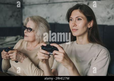 Frau mit ihrer leitenden Mutter, die Videospiele auf einer Spielkonsole im Wohnzimmer spielt. Zwei Generationen spielender Joystick. Stockfoto