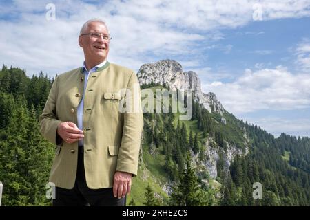 Aschau Im Chiemgau, Deutschland. 16. Aug. 2023. Joachim Herrmann (CSU) Staatsminister für Inneres, Sport und Integration steht während einer Ernennung vor dem Panorama des Kampenwand. Herrmann informierte sich über die Ernennung zu den Themen alpenunfallstatistik, "sicheres Mountainbiken" und "Sicherheitsausrüstung für Via ferrata". Auch die Demonstration einer Bergrettung mit einem Helikopter, die als Einsatzmöglichkeit in den Bayerischen Bergen unverzichtbar ist, wurde gezeigt. Kredit: Peter Kneffel/dpa/Alamy Live News Stockfoto