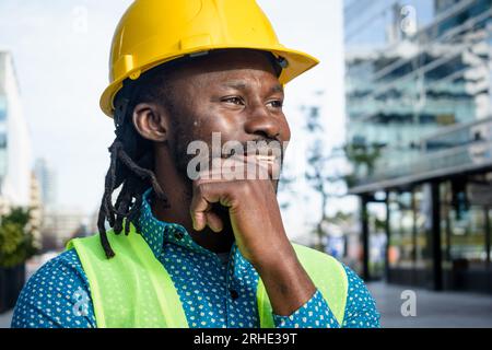 Afrikanischer Bauingenieur, der Sicherheitshelm trägt, im Freien steht, sehr selbstbewusst lächelt und sich lächelnd umschaut, Polizist Stockfoto