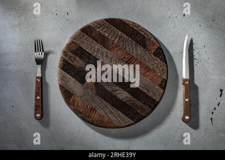 Rundes Holzschneidebrett mit Gabel und Messer für Steak auf rauer Betonkulisse. Hintergrund für Ihren Kopierbereich oder Ihr Design Stockfoto