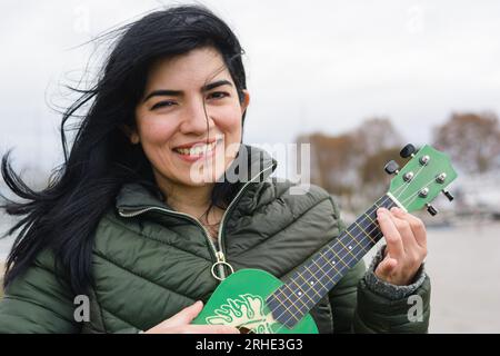 Junge venezolanische Frau, glücklich spielend Ukulele spielen lernend, stehend, Kamera schauend, in argentinien draußen reisen, Lifestyle-Konzept, Copy-Spac Stockfoto