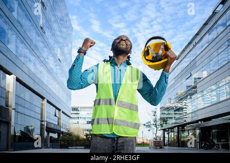 Bauingenieur Mann afrikanischer Herkunft lächelt sehr erfolgreich und glücklich, mit geballter Faust und hält seinen Helm mit einer Hand und feiert Tag an Stockfoto