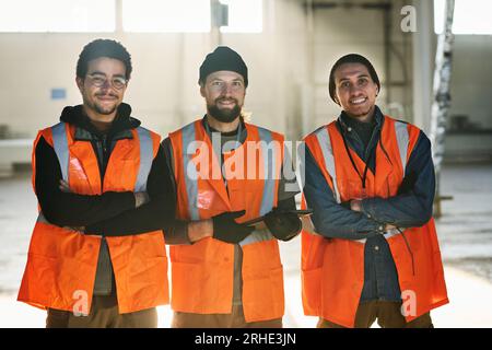 Drei junge, erfolgreiche Ingenieure aus der Fabrik oder Arbeiter aus dem Lager in Sicherheitswesten, die ihre Arme an der Brust kreuzen und dich ansehen Stockfoto