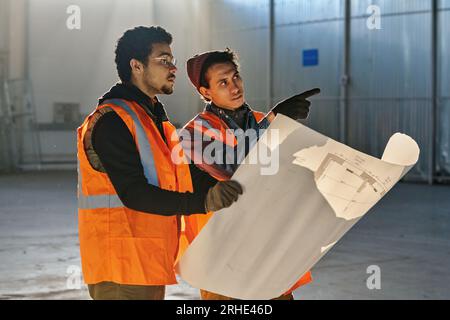 Einer von zwei Ingenieuren zeigte nach vorne, während er seinem Kollegen, der bei der Besprechung im Lager einen großen Entwurf abhielt, Skizzenpunkte erläuterte Stockfoto