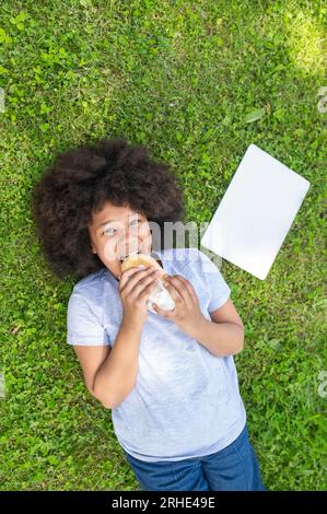 Ein Mädchen, das auf dem Gras liegt und Fast Food im Park isst, ein glückliches Kind, das einen Snack hat, nachdem es seinen Laptop im Freien benutzt hat. Stockfoto