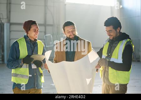 Glücklicher junger Bauunternehmer und zwei Mitarbeiter in Sicherheitsjacken, die sich einen Entwurf oder einen Bauplan auf einem Bauplan ansehen, während sie vor der Kamera stehen Stockfoto