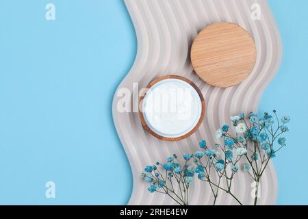 Kosmetische cremefarbene Dose auf Beton Dekorationswelle mit Feldblumen, minimalistisches Design auf blauem Hintergrund, Draufsicht Stockfoto