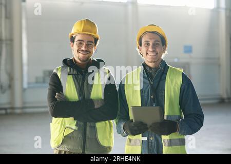 Zwei fröhliche, junge, multiethnische Ingenieure in Schutzhelmen und reflektierenden Jacken stehen vor der Kamera im Lager eines Industriewerks oder einer Fabrik Stockfoto