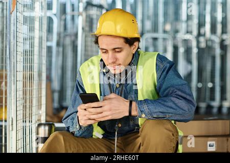 Junger männlicher Ingenieur in Arbeitskleidung und Schutzhelm, der SMS auf einem Smartphone schreibt, während er auf einem großen metallischen Industriewagen mit Boxen vor der Kamera sitzt Stockfoto