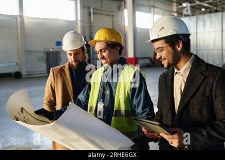 Seitenansicht dreier junger erfolgreicher Architekten, die Skizzenpunkte zum Entwurf diskutieren, den der Mensch in Schutzhelm und Sicherheitsjacke trägt Stockfoto
