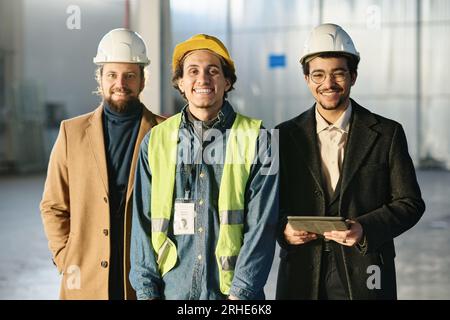 Drei junge, erfolgreiche Ingenieure in Schutzhelmen stehen vor der Kamera und schauen Sie an, während einer von ihnen ein Tablet in der Hand hält Stockfoto