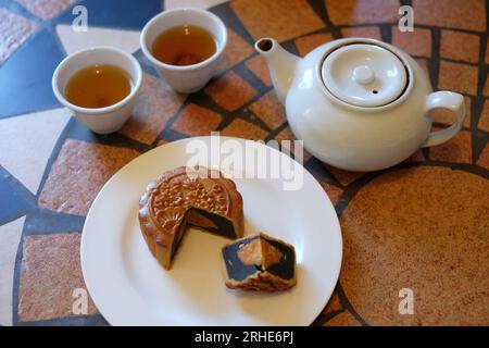 Chinesischer Mondkuchen mit einem Viertel entfernt und zeigt das runde Eigelb in seiner dunklen Bohnenpastenfüllung, zwei Teetassen und eine Teekanne auf einem Mosaiktisch. Stockfoto