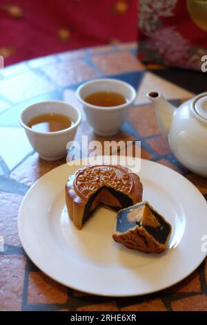 Chinesischer Mondkuchen mit einem Viertel entfernt und zeigt das runde Eigelb in seiner dunklen Bohnenpaste, zwei Teetassen und eine Teekanne auf einem Mosaiktisch. Stockfoto