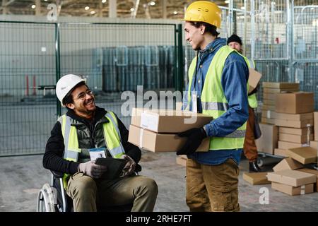 Glücklicher junger Ingenieur im Rollstuhl, der einen Kollegen beobachtet, der einen Stapel verpackter Pappkartons trägt, während er mit ihm während der Arbeit im Lager spricht Stockfoto
