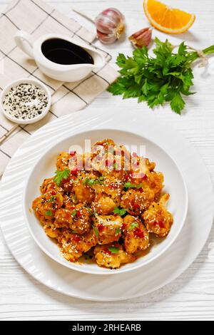 Knusprige Blumenkohlblüten mit Sesamsamen, Chiliflocken und Petersilie in weißer Schüssel auf weißem Holztisch mit Zutaten, vertikal Stockfoto