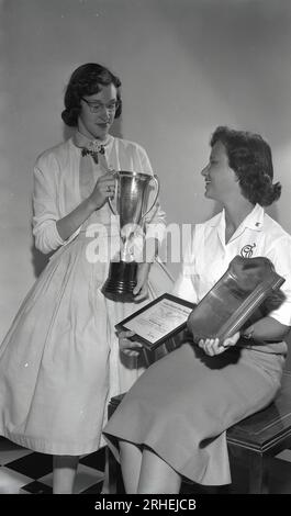 1954, historisch, zwei junge Damen, High School Schüler, eine steht mit einer Trophäe, die andere sitzt mit ihren Auszeichnungen, eine Plakette und ein gerahmtes Zertifikat, mit einer First Class Honour von der National Scholastic Press Association (NSPA), USA. Die NSPA, eine gemeinnützige Organisation, wurde 1921 für den Journalismus in Veröffentlichungen von Highschool- und Sekundarschulen gegründet und gilt als eine der angesehensten Preisverleihungsgremien. Die junge Dame hier mit der Tafel hat den All American Award gewonnen. Stockfoto