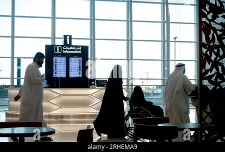 Flughafen Bahrain. Passagiere, die in einer Silhouette vorbeifahren, mit Hintergrundbeleuchtung. Informationsschalter. Frauen im Hijab mit Rollstuhl. Männer in weißer Kleidung Stockfoto