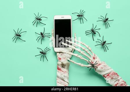 Skeletthände mit modernem Mobiltelefon und Spinnen für die Halloween-Feier auf türkisfarbenem Hintergrund Stockfoto