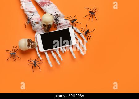 Skeletthände mit modernem Mobiltelefon, Spinnen und Schädel für die Halloween-Feier auf orangefarbenem Hintergrund Stockfoto