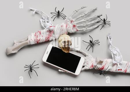 Skeletthände mit modernem Handy, Schädel und Spinnen für die Halloween-Feier auf grauem Hintergrund Stockfoto