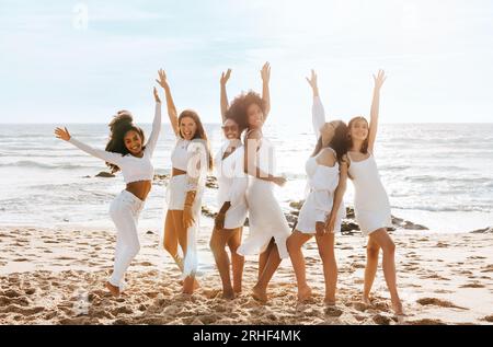 Glückliche, multirassische Damen tanzen mit erhobenen Armen, während sie am sonnigen Tag eine Junggesellinnenparty am Strand feiern Stockfoto