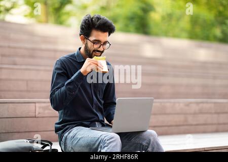 Lachender indianer, der Sandwich aß, sich mit Laptop im Park entspannte Stockfoto