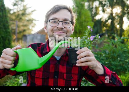 Der junge, lächelnde Blumenhändler in Gläsern hält eine grüne Gießkanne und eine Pflanze im Freien Stockfoto