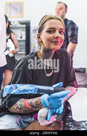 Lächelnde, positive Tätowierungskünstlerin, die beim Tätowieren in die Kamera schaut. Verschwommener Hintergrund. Tätowierende Studiowerbung. Vertikale Innenaufnahme. Hochwertige Fotos Stockfoto