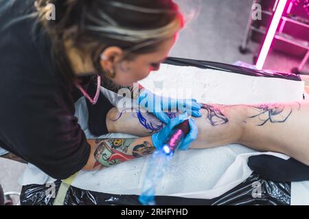 Eine Tätowierin, die ein Tattoo auf das Bein eines Klienten zeichnet. Konzeptkunst Hochwinkelaufnahme im Innenbereich. Verschwommener Hintergrund. Hochwertige Fotos Stockfoto