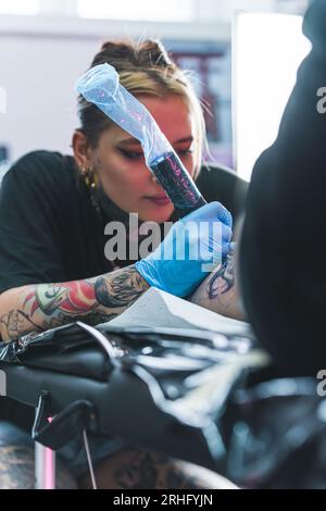 Fokussierte Tätowierungskünstlerin, die ein Tattoo auf ihre Klientin zeichnet. Beintattookonzept. Echte Menschen, die Kunst erschaffen Vertikale Innenaufnahme. Hochwertige Fotos Stockfoto