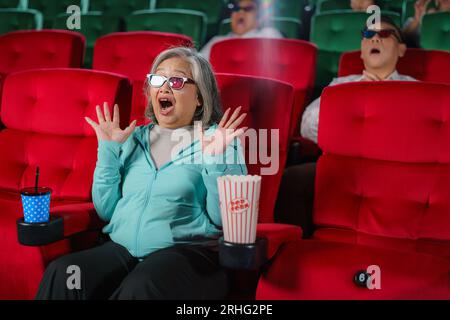 Ältere asiatische Frauen mit Brille sehen sich gern 3D-Filme und Popcorn in der Hand an und genießen das faszinierende Erlebnis. Stockfoto