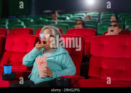 Ältere asiatische Frauen mit Brille sehen sich gern 3D-Filme und Popcorn in der Hand an und genießen das faszinierende Erlebnis. Stockfoto