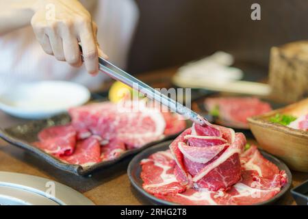 Die Hand einer Frau verwendet Zangen, um Wagyu-Rindfleisch auf einen Teller zu legen, bereit zum Grillen über Holzkohle, was das kulinarische Erlebnis in einem japanischen Restaurant bereichert Stockfoto