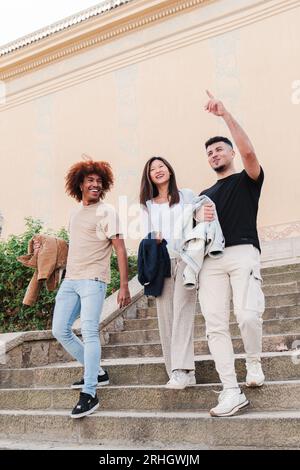 Vertikales Porträt einer Gruppe von multiethnischen Studenten, die unten auf dem Campus zusammenlaufen, reden, zeigen und lächeln. Junge Teenager-Frau mit zwei männlichen Freunden, die auf einer Reise auf Sightseeing gehen. Hochwertige Fotos Stockfoto