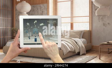 Smart Remote Home Control System auf einem digitalen Tablet. Gerät mit App-Symbolen. Modernes japandi-Schlafzimmer mit Doppelbett und Tapete im Hintergrund, Stockfoto