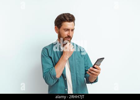 Nachdenklicher Mann, der auf sein Handy schaut und seinen Bart berührt, der auf weißem Hintergrund steht Stockfoto
