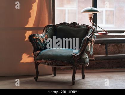 Klassischer Sessel im Vintage-Stil mit schönen geschnitzten Holzelementen und blau zerrissenen Polstern im Grunge-Raum mit grüner Lampe auf der Fensterbank. Retro-Innenkonzept mit Kopierraum. Stockfoto