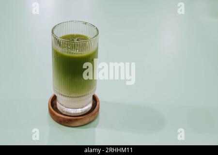 Selektiver Fokusschuss eines Glases Ice Matcha oder japanischer grüner Tee Latte auf Holzuntersetzer auf isoliertem salbeigrünen Pastell-Tisch Stockfoto