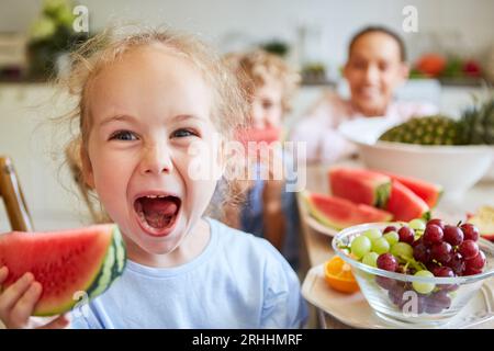 Blondes, verspieltes Mädchen, das zu Hause mit der Familie im Hintergrund schreit, während es frische Wassermelonenscheiben hält Stockfoto