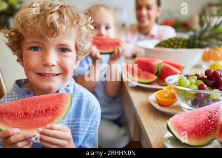 Lächelnder Junge, der Wassermelonenscheiben mit frischem Obst auf dem Tisch in der Küche zu Hause hält Stockfoto
