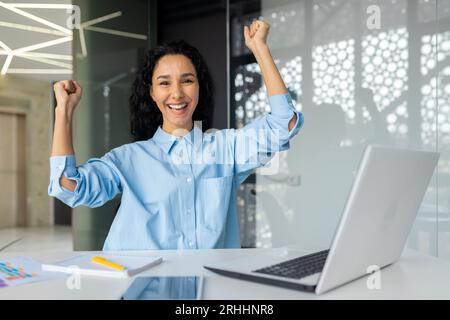 Porträt einer verschlafenen, Reifen Geschäftsfrau, einer lateinamerikanischen Gewinnerin, die die Hände hoch hält, die Hand hoch hält, die Hand hoch hält und im Büro mit einem Laptop arbeitet. Stockfoto