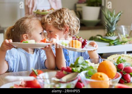 Verspielte Geschwister haben Spaß, während sie Obstsalatplatten in der Küche zu Hause halten Stockfoto