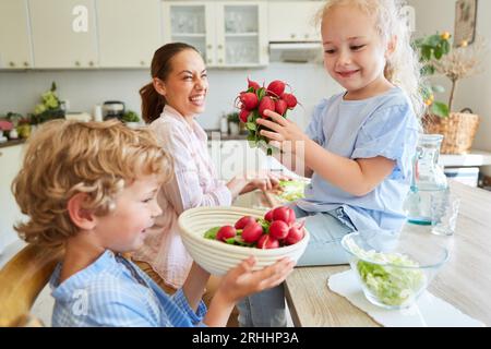 Lächelndes Mädchen, das dem Bruder hilft, eine Schale Rettich zu halten, während er mit der Mutter zu Hause in der Küche kocht Stockfoto