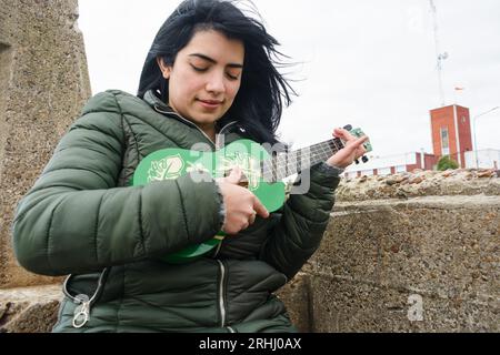 Die junge Latina aus venezolanischer Herkunft in der Winterjacke sitzt draußen an einem kalten und bewölkten Tag und lernt Ukulele zu spielen, Kopierraum. Stockfoto