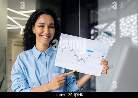 Eine Geschäftsfrau schaut bei einem Online-Meeting in die Laptop-Kamera und zeigt ihren Kollegen und Partnern eine Grafik des finanziellen Einkommens, eine zufriedene Finanzierin bei einer Startup-Präsentation im Büro Stockfoto