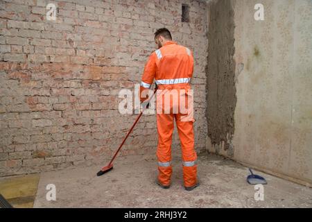 Erbauer reinigt den Raum nach Abbrucharbeiten von Staub und trägt Handschuhe, Stiefel und orangefarbene Overalls. Stockfoto